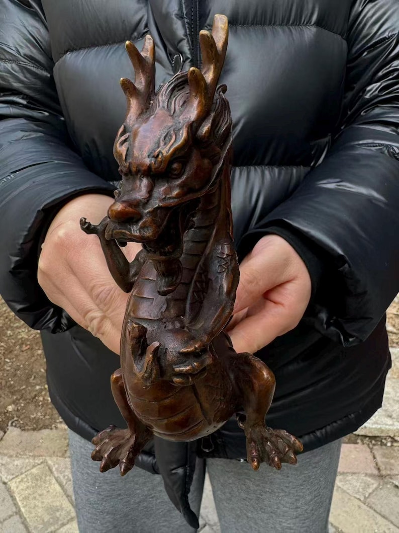 K&ouml;nnte beinhalten: Bronze-Drachenfigur mit aufwendigen Details. Der Drache hat ein detailliertes Gesicht, Geweihe und Schuppen. Er ist mit verschr&auml;nkten Armen und F&uuml;&szlig;en auf dem Boden posiert. Die Figur ist ein dekorativer Gegenstand.