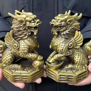 May include: Two antique brass-colored mythical creatures, possibly dragons or lions, are displayed. Each figure has wings, a detailed mane, and is seated on a square base. One holds a small bowl, the other a small ball. The figures are facing each other.