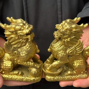 May include: Two gold-colored, detailed statues of mythical creatures, possibly dragons or lions, are held in hands. The statues have intricate designs, including wings, scales, and ornate features. They are displayed against a dark background.