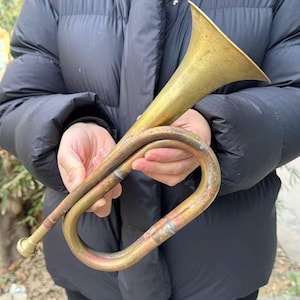 Può includere: Una tromba vintage in ottone con campana svasata e tubi curvi. La tromba presenta segni di età e utilizzo, con un aspetto ossidato. Lo strumento è tenuto su uno sfondo scuro.