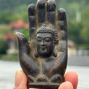 May include: A bronze-colored Buddha statue in the shape of a hand. The Buddha's face is centered in the palm, with detailed facial features. The hand has a textured surface and is held up against a blurred outdoor background.