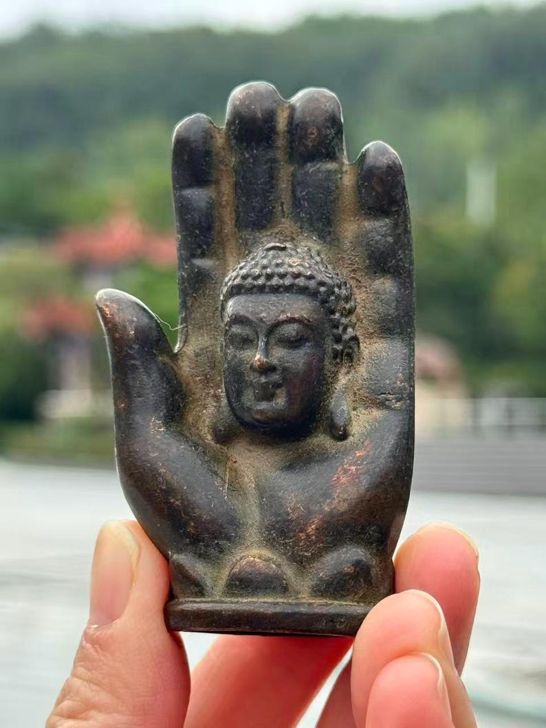 May include: A bronze-colored Buddha statue in the shape of a hand. The Buddha's face is centered in the palm, with detailed facial features. The hand has a textured surface and is held up against a blurred outdoor background.