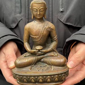 以下が含まれることがあります： 座禅の姿勢のブロンズ仏像。穏やかな表情、詳細なローブがあり、装飾的な台座に座っています。像は手で持たれています。