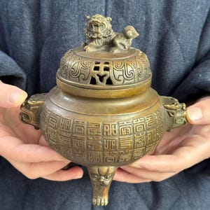 May include: A bronze-toned metal incense burner featuring a lid topped with a sculpted lion figure. The body is decorated with a grid of embossed Chinese characters and side handles shaped like animal heads, supported by three ornate legs.