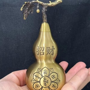 May include: A brass-colored metal gourd-shaped vessel featuring a textured stem with a small hanging charm. The upper section displays the Chinese characters 招财, while the lower section features a circular lotus-style mandala with Sanskrit script.