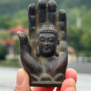 May include: A bronze-colored Buddha statue in the shape of a hand. The Buddha's face is centered in the palm, with detailed facial features and a serene expression. The hand's fingers are slightly curved, and the overall design is intricate.