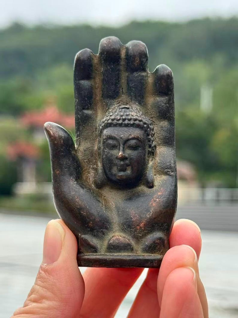 May include: A bronze-colored Buddha statue in the shape of a hand. The Buddha's face is centered in the palm, with detailed facial features and a serene expression. The hand's fingers are slightly curved, and the overall design is intricate.