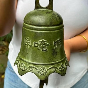 Op de afbeelding: Een groene metalen bel met Chinese karakters op de voorkant. De bel heeft een decoratief bloemmotief langs de onderrand en een lus aan de bovenkant om op te hangen. De bel is opgehangen aan een donkere metalen haak.