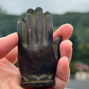 May include: A dark bronze-colored hand sculpture with detailed fingers and a decorative base. The sculpture is held in a hand, showcasing its size and texture. The hand is in a gesture of blessing or offering.