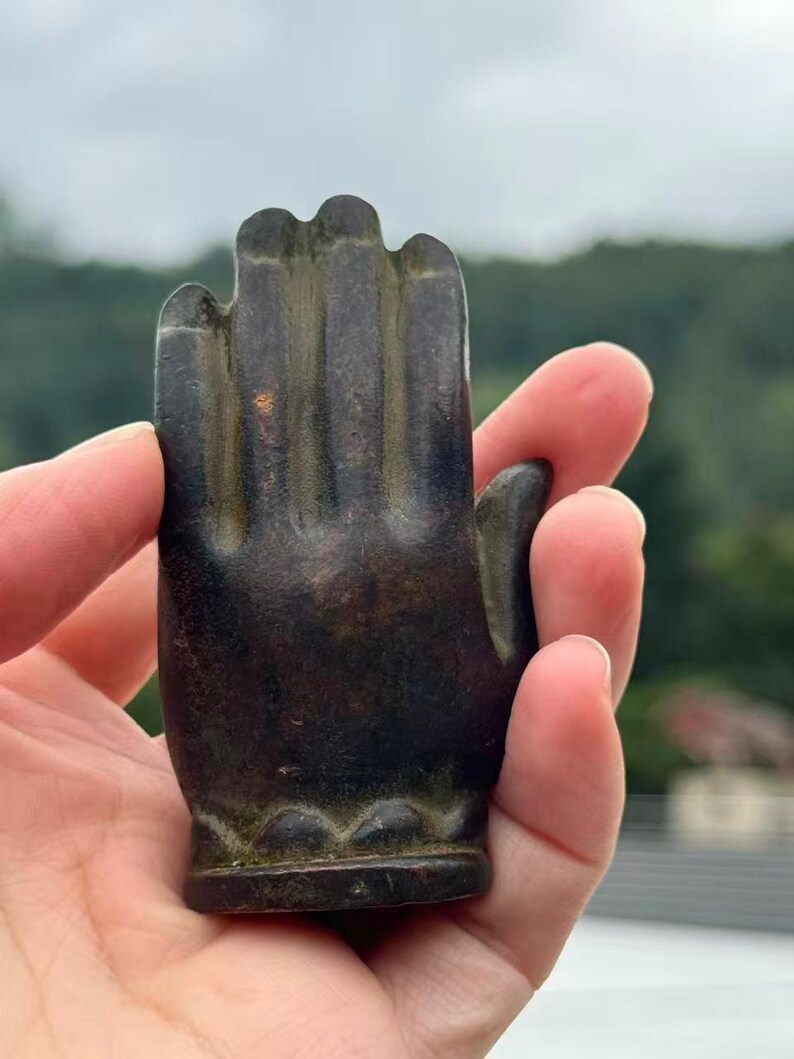 May include: A dark bronze-colored hand sculpture with detailed fingers and a decorative base. The sculpture is held in a hand, showcasing its size and texture. The hand is in a gesture of blessing or offering.