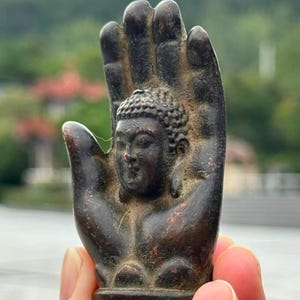 May include: A bronze-colored Buddha statue within a hand-shaped sculpture. The Buddha's face is detailed with a serene expression. The sculpture is held up against a blurred natural background.
