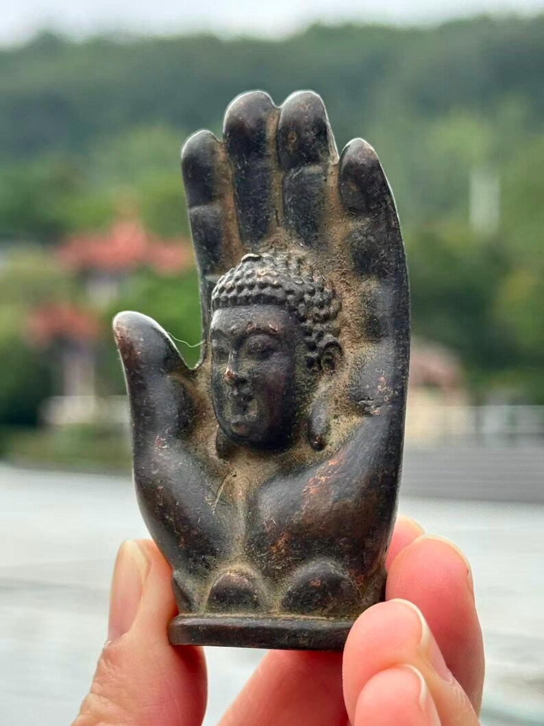 May include: A bronze-colored Buddha statue within a hand-shaped sculpture. The Buddha's face is detailed with a serene expression. The sculpture is held up against a blurred natural background.