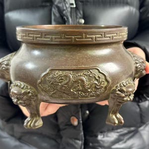 May include: A bronze-toned metal incense burner featuring a wide, rounded body with a relief carving of a dragon on the front. The rim is decorated with a geometric Greek key pattern, and the vessel is supported by three legs shaped like lion heads.
