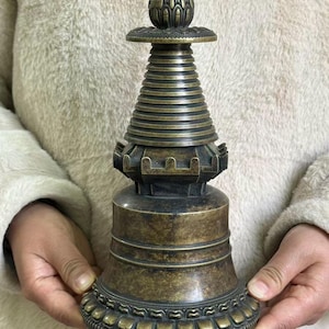 May include: A bronze-colored metal pagoda sculpture featuring a tiered conical spire, a square mid-section with notched edges, and a wide, bell-shaped base. The bottom edge is decorated with a repeating petal pattern, and the top is capped with a finial.