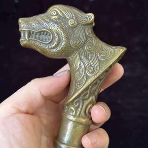 Pode incluir: Um punho de bengala em metal cor de latão com a forma de uma cabeça de lobo ou canídeo estilizada. A superfície apresenta padrões gravados intrincados e em espiral ao longo do pescoço e focinho, com uma base cilíndrica para fixação.