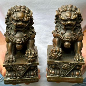 May include: Two bronze-colored Foo Dog statues. Each statue is in a seated position on a rectangular base with ornate carvings. The Foo Dogs have detailed manes, fierce expressions, and are holding a ball under their paws. The statues are approximately 5 inches tall.