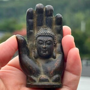 May include: A bronze-colored Buddha statue is set within a hand-shaped sculpture. The Buddha figure is centered, with a serene expression and detailed hair. The hand's fingers are slightly curved, and the overall design has an antique appearance.