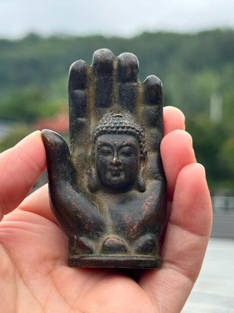 May include: A bronze-colored Buddha statue is set within a hand-shaped sculpture. The Buddha figure is centered, with a serene expression and detailed hair. The hand's fingers are slightly curved, and the overall design has an antique appearance.