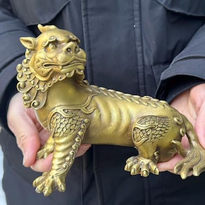 May include: A golden-colored, detailed sculpture of a mythical creature, possibly a guardian lion. The statue has intricate scales, a flowing mane, and a fierce expression. The sculpture is held in both hands.