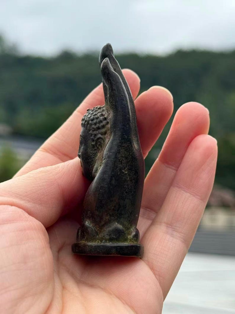 May include: A small, dark bronze-colored Buddha statue, featuring a detailed head and raised hand. The statue has a textured surface and is mounted on a small, rectangular base. The sculpture is held in a hand.
