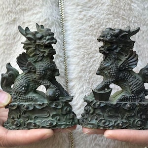 May include: Two antique bronze-colored statues of mythical creatures. Each statue features a detailed dragon-like figure with wings, perched on a cloud-like base. One holds a small object. The statues are held in hands.