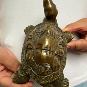 May include: A bronze turtle figurine with a detailed shell pattern and outstretched head. The sculpture has a weathered appearance, showcasing the intricate design of the shell and the turtle's form. The figurine is held in a person's hands.