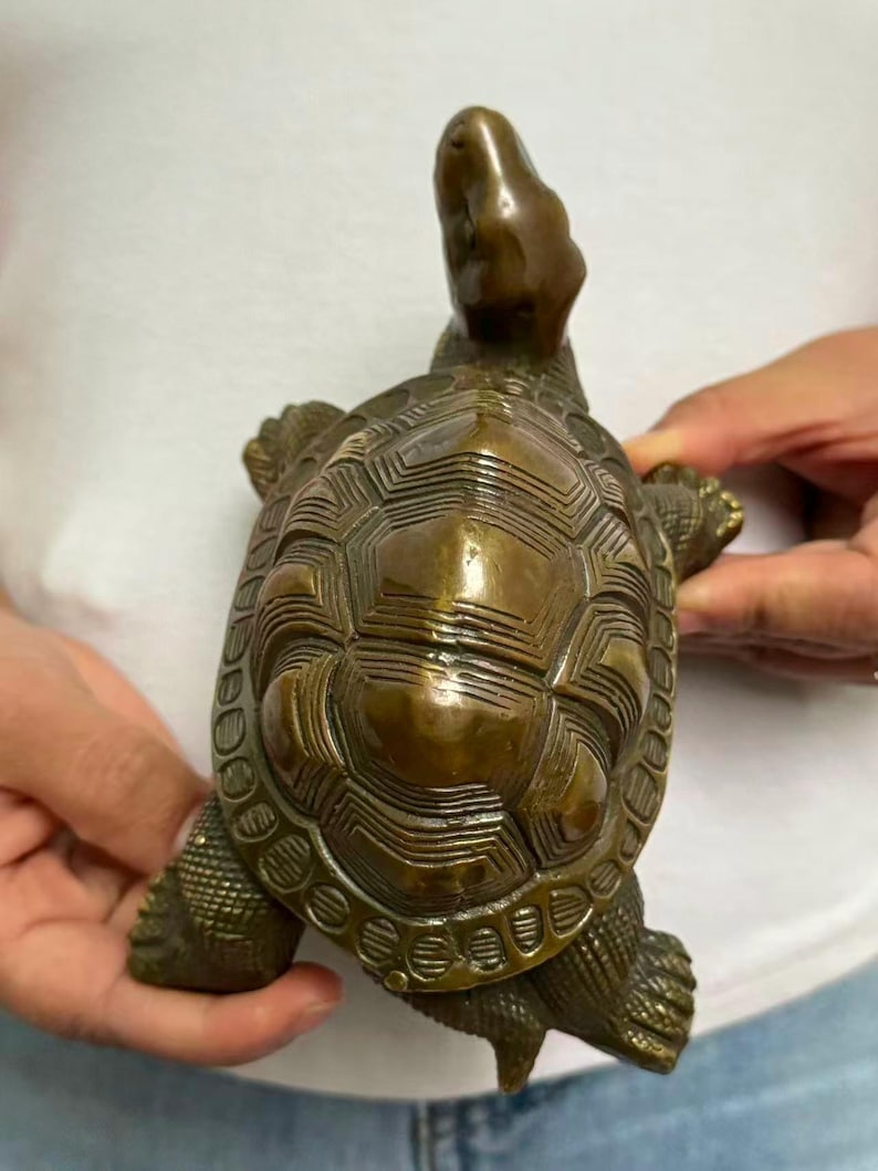 May include: A bronze turtle figurine with a detailed shell pattern and outstretched head. The sculpture has a weathered appearance, showcasing the intricate design of the shell and the turtle's form. The figurine is held in a person's hands.