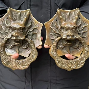 May include: Two antique bronze door knockers. Each knocker features a detailed dragon head with an open mouth holding a decorative ring. The dragon heads are mounted on a shield-shaped backplate. The bronze has a weathered patina.