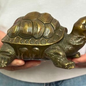 May include: A bronze-colored turtle figurine with a detailed shell pattern and textured skin. The sculpture is held in hands, showcasing its three-dimensional form and realistic features. The turtle's head is raised, and its legs are extended.