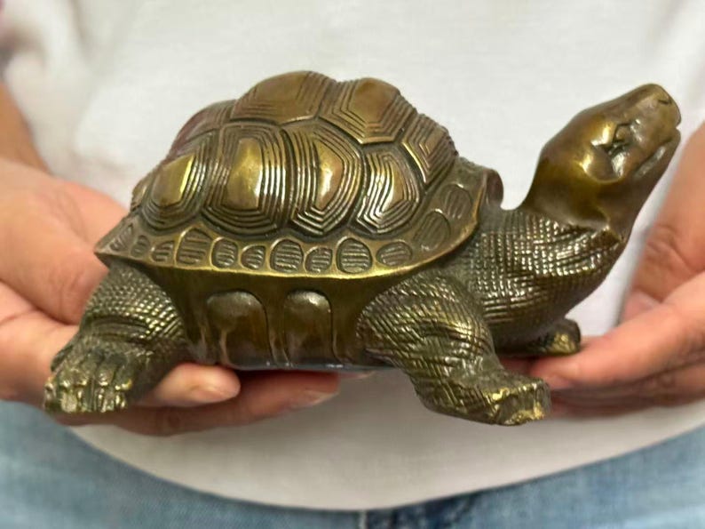May include: A bronze-colored turtle figurine with a detailed shell pattern and textured skin. The sculpture is held in hands, showcasing its three-dimensional form and realistic features. The turtle's head is raised, and its legs are extended.