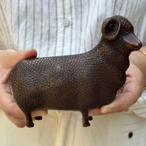 May include: A bronze-toned metal figurine of a ram standing in profile. The sculpture features a textured, curly wool pattern covering the body, a smooth face, and curved horns. The figure is depicted in a standing position with four legs.