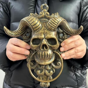 May include: A bronze-colored skull-shaped door knocker with large horns and a ring handle. The knocker has intricate details and a weathered finish, giving it a vintage appearance. The skull features detailed eye sockets, a nose cavity, and a row of teeth.