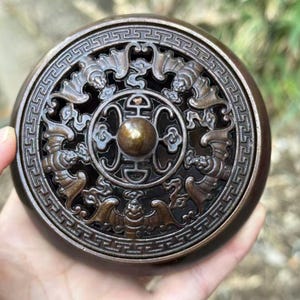 May include: A round, bronze-colored incense burner with intricate relief carvings. The lid features a central design with a raised knob and is surrounded by a detailed border with bat and geometric patterns. The burner is held in a hand.