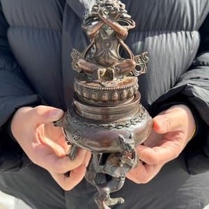 May include: Bronze-colored incense burner with a seated figure in a meditative pose. The burner features intricate details, including a lotus base and animal-like figures. The figure's hands are crossed over its mouth.