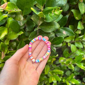 May include: A colorful bracelet made of small, round, flat beads. The beads are arranged in a striped pattern of pink, yellow, blue, and white.