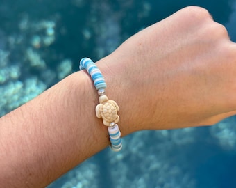 Könnte beinhalten: Ein blaues und weiß gestreiftes Armband mit einem kleinen weißen Schildkrötenanhänger. Das Armband wird am Handgelenk einer Person vor einem blauen Hintergrund getragen.