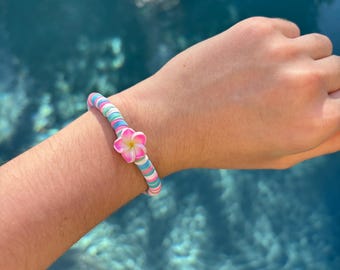 May include: A colorful beaded bracelet with a pink flower charm. The bracelet is made of small, round beads in shades of blue, pink, and white. The flower charm is a single pink flower with white petals.
