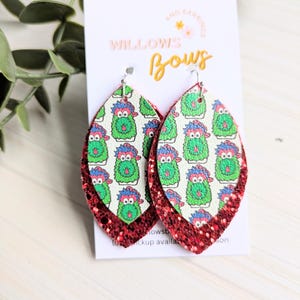 May include: A pair of layered, teardrop-shaped earrings. The top layer features a repeating pattern of green monster faces on a white background. The bottom layer is a red glitter material. The earrings are displayed on a white card with the text "WILLOWS Bows".