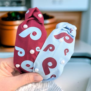 May include: Two fabric headbands with a swirl pattern and pearl embellishments. One headband is burgundy with white swirls and the other is light blue with maroon swirls.