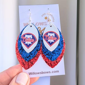 May include: A pair of red, white, and blue glitter earrings with a Phillies logo. The earrings are shaped like a teardrop and have a silver hook.