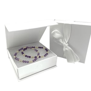 May include: Two amethyst bead bracelets with gold-colored beads are displayed inside a white gift box. The box is open, revealing the jewelry on a bed of white material. A second white box with a tied white ribbon is visible.