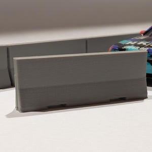 May include: A gray, rectangular model barrier with horizontal lines, resembling a concrete barrier. A colorful toy car is partially visible in the background. The barrier is designed for model car displays or dioramas.