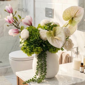 May include: A white ceramic vase holding a floral arrangement. The arrangement features pink magnolia branches, white roses, green hydrangeas, and white anthurium flowers. The vase is on a marble countertop in a bathroom.