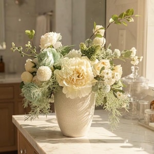 K&ouml;nnte beinhalten: Ein Blumenarrangement in einer strukturierten, cremefarbenen Vase. Der Strau&szlig; besteht aus gro&szlig;en, cremefarbenen Pfingstrosen, zarten blau-gr&uuml;nen Akzenten und verschiedenen wei&szlig;en und gr&uuml;nen Bl&auml;ttern. Die Anordnung steht auf einer Marmorarbeitsplatte.