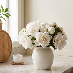 Op de afbeelding: Een boeket witte pioenrozen in een matte witte keramische vaas met horizontale ribbels. De bloemen hebben gelaagde bloemblaadjes en groene bladeren, wat zorgt voor een vol, rond bloemstuk in een eenvoudig, neutraal gekleurd vaasje.