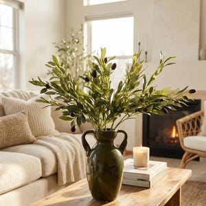 May include: A green ceramic vase with two handles holds olive branches with green leaves and black olives. The vase sits on a wooden coffee table in a living room setting. A lit candle and books are also on the table.