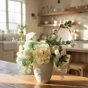 K&ouml;nnte beinhalten: Ein Blumenarrangement in einer strukturierten, hellfarbigen Vase. Der Strau&szlig; zeigt gro&szlig;e, cremefarbene gelbe Pfingstrosen, wei&szlig;e und hellgr&uuml;ne Bl&uuml;ten und verschiedene gr&uuml;ne Bl&auml;tter. Die Vase steht auf einem Holztisch in einem hellen Raum.