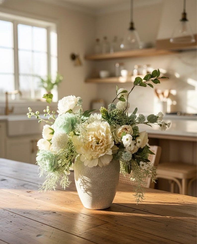 K&ouml;nnte beinhalten: Ein Blumenarrangement in einer strukturierten, hellfarbigen Vase. Der Strau&szlig; zeigt gro&szlig;e, cremefarbene gelbe Pfingstrosen, wei&szlig;e und hellgr&uuml;ne Bl&uuml;ten und verschiedene gr&uuml;ne Bl&auml;tter. Die Vase steht auf einem Holztisch in einem hellen Raum.