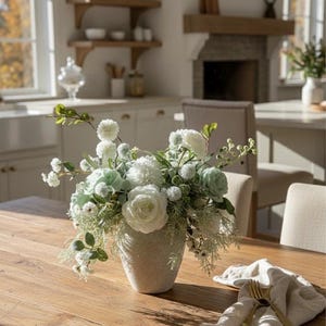 K&ouml;nnte beinhalten: Ein Blumenarrangement mit wei&szlig;en und hellgr&uuml;nen Bl&uuml;ten in einer strukturierten, hellfarbigen Vase auf einem Holztisch. Auf dem Tisch liegt eine gefaltete Leinen-Serviette mit goldenem Besteck.
