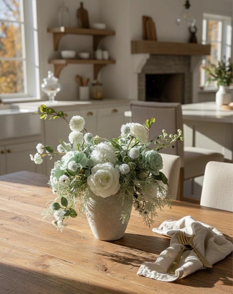 K&ouml;nnte beinhalten: Ein Blumenarrangement mit wei&szlig;en und hellgr&uuml;nen Bl&uuml;ten in einer strukturierten, hellfarbigen Vase auf einem Holztisch. Auf dem Tisch liegt eine gefaltete Leinen-Serviette mit goldenem Besteck.
